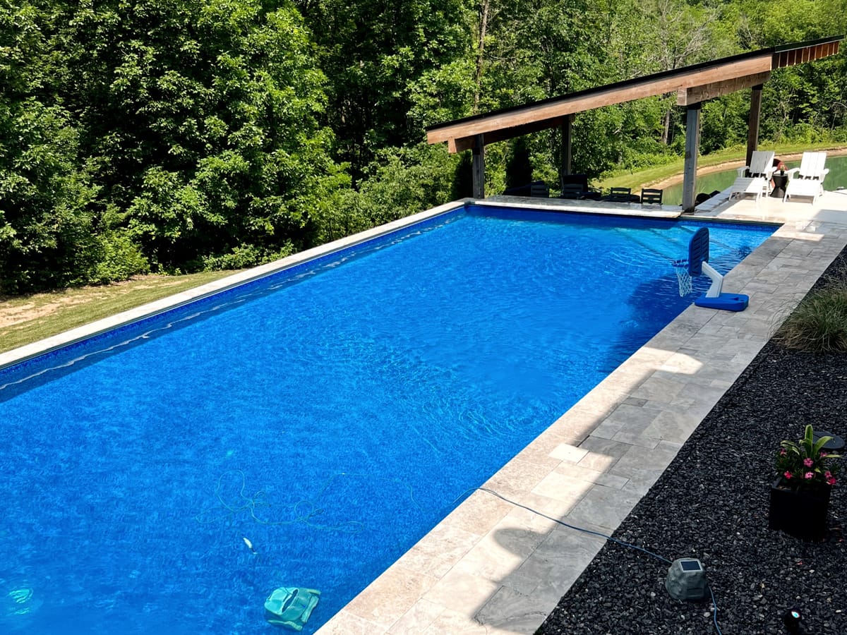 Long rectangular swimming pool with a pergola and dark coping beside a wooded backyard